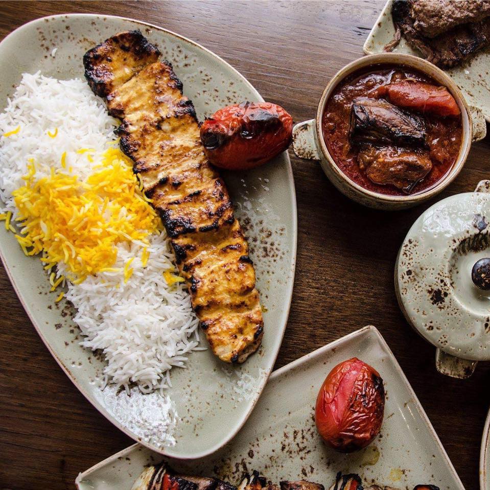 Grilled chicken kabob next to saffron rice and a grilled tomato on an oval plate. Surrounding items include a bowl of stew and grilled tomatoes on smaller plates, all on a wooden table