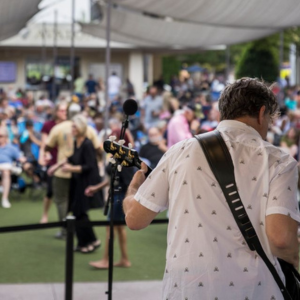 A musician plays guitar on an outdoor stage, facing a large, seated audience under a canopy, with people mingling and watching the performance