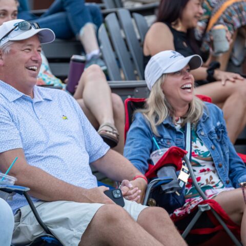 A man and woman, wearing casual clothing and white caps, laugh while sitting on folding chairs They hold hands, surrounded by other seated people in an outdoor setting