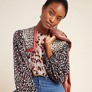 A person wearing a colorful, patterned blouse and blue jeans slightly tilts their head, holding a rust-colored jacket over their shoulder, posed against a neutral background