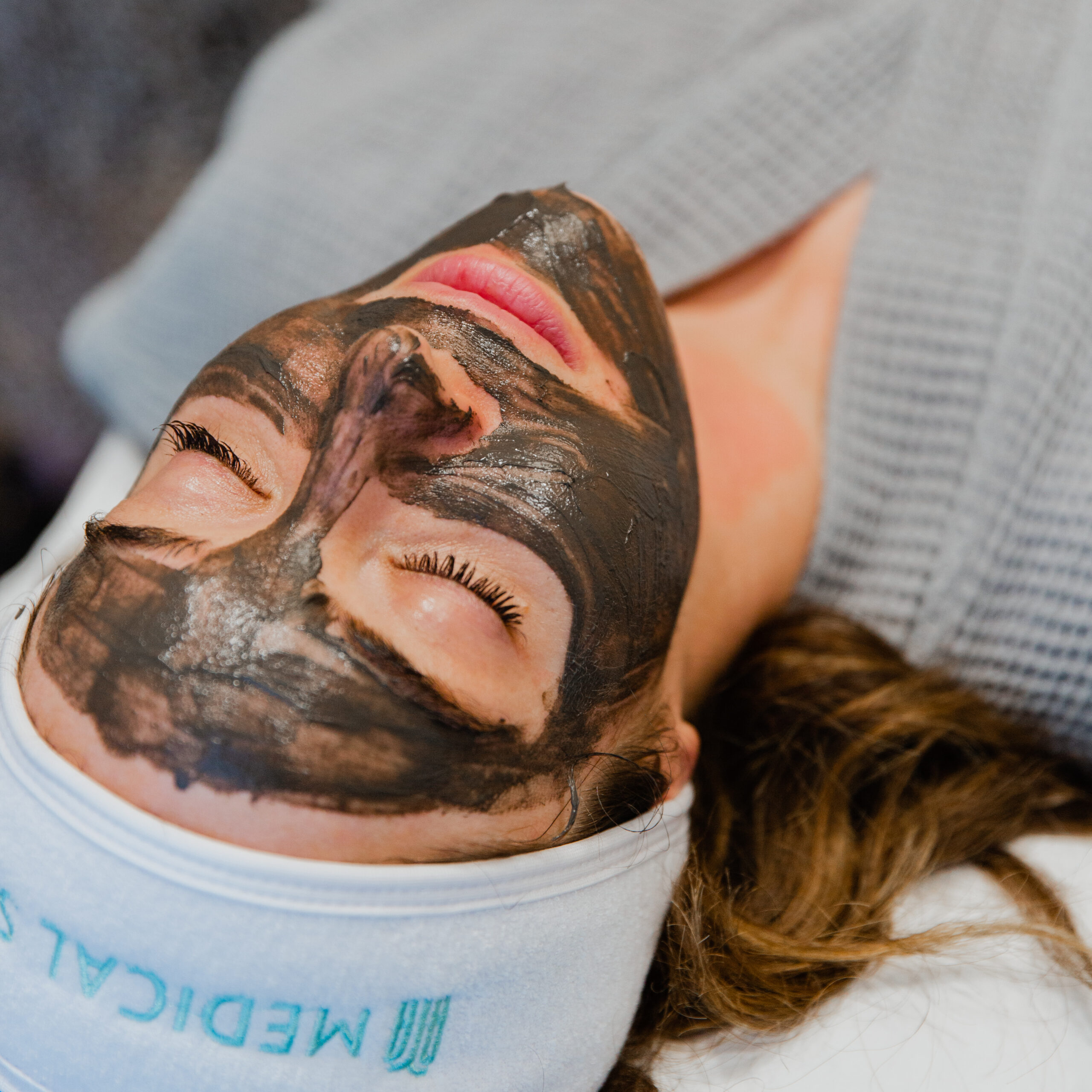 A person lies down with a dark facial mask applied, wearing a headband that says 