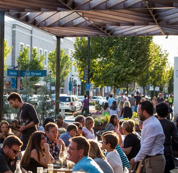 A group of people eating and drinking at an outdoor restaurant under a wooden canopy on a sunny day in a bustling urban area with leafy trees and street signs reading