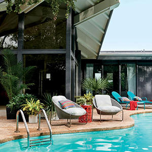 Two cushioned lounge chairs sit by the edge of a curved swimming pool, accompanied by plants and another pair of blue lounge chairs, outside a modern glass-and-metal building