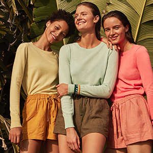 Three smiling individuals in pastel-colored sweaters and shorts stand closely together against a backdrop of large green leaves, suggesting a tropical environment