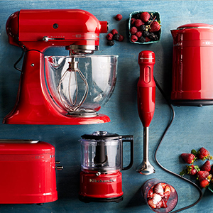 Red kitchen appliances including a stand mixer, blender, toaster, and kettle are arranged on a blue countertop along with scattered strawberries and a bowl of sliced strawberries
