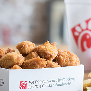 Fried chicken nuggets sit in a white box labeled “We Didn’t Invent The Chicken, Just The Chicken Sandwich” with the Chick-fil-A logo A drink cup with the same logo appears in the background