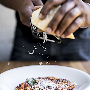 Hands grate cheese over a bowl of pasta, situated on a wooden table with a blurred background Thin strands of cheese are visibly falling onto the pasta