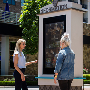 Two women interact with an outdoor information kiosk featuring a screen displaying various advertisements. They stand on a paved area with buildings, greenery, and sunlight surrounding them