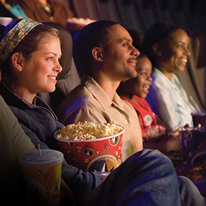 People sit in a movie theater, holding drinks and popcorn, intently watching the screen, smiling and appearing engaged with the film