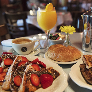 A table set for breakfast with French toast topped with strawberries, a croissant, coffee, orange juice, and a yellow flower in a vase, in a cozy café