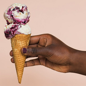 A hand holds a waffle cone with two scoops of berry-swirled vanilla ice cream against a plain beige background