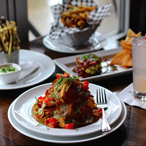 A plate of crispy fries topped with chopped vegetables sits on a wooden table, surrounded by other food dishes, a drink, and additional condiments in a restaurant setting