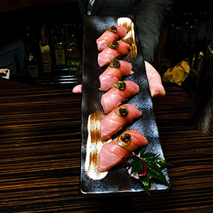 A person holds a rectangular black plate with six pieces of sushi topped with garnish, against a dark wood-grain table in a dimly lit bar with bottles in the background