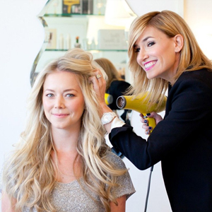 A hairstylist blow-dries a seated woman's long hair in a salon, with shelves of hair products and a decorative mirror in the background