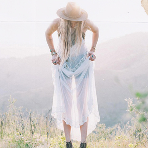 A person wearing a hat and a sheer dress looks down, slightly bent forward; they are outdoors in a grassy, hilly landscape with a light, hazy sky