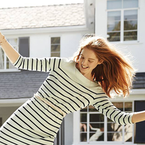 A woman wearing a striped dress is joyfully dancing with arms outstretched in front of a white house with large windows