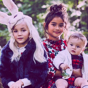 Three children sit in a forest clearing, dressed in formal attire. The child on the left holds a white mask, the middle child wears a rabbit headpiece, and the right child wears floral patterns