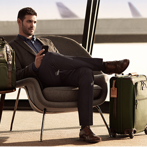 A man in a suit sits in an airport lounge chair, holding a smartphone with crossed legs Green luggage is beside him, and airplane tails are visible outside the window
