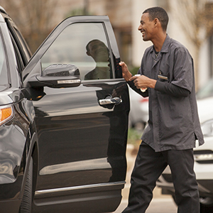 A person in a gray uniform opens the driver's door of a black SUV in a parking lot, smiling and holding keys, with other parked cars in the background
