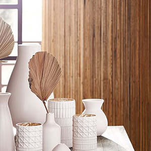 White ceramic vases with geometric textures arranged on a marble surface, containing dried palm leaves, set against a vertical wooden panel background with sunlight filtering through a window