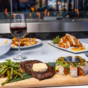 A grilled steak topped with butter, green beans, sushi, and a cup of sauce sit on a wooden board; a glass of red wine and additional dishes in a restaurant setting