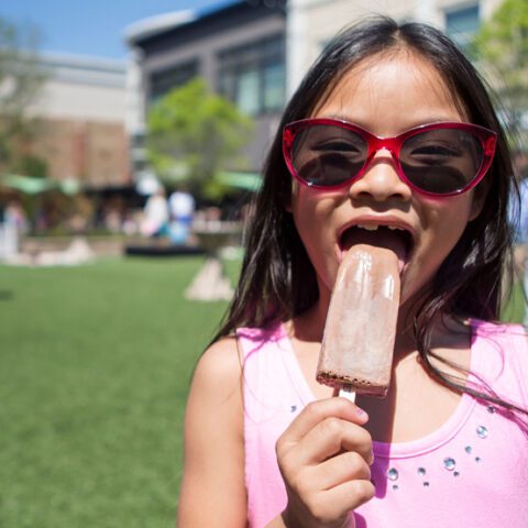 Child with red sunglasses eating a popsicle in a sunny outdoor plaza with green grass, buildings, and trees in the background
