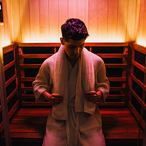 A man wearing a white robe sits in a warmly lit wooden sauna, looking down and holding the robe's lapels. The sauna interior features wooden benches and paneled walls