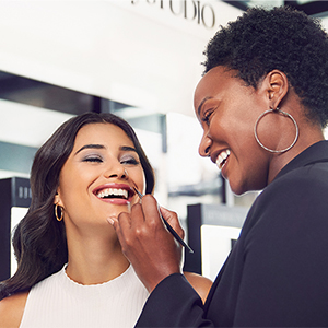 A smiling woman is holding a makeup brush and applying lip color to another smiling woman in a well-lit professional studio