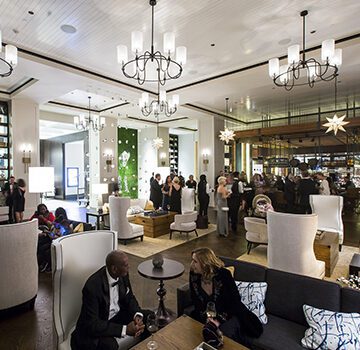 People converse and socialize in a well-lit, elegantly decorated lounge. White chandeliers hang from the ceiling, and patrons are seated on comfortable chairs and couches, with a bar in the background
