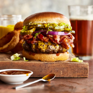 A cheeseburger with bacon, lettuce, pickles, and onion sits on a wooden board, accompanied by onion rings, mustard, a glass of beer, and a dish of sauce with a spoon