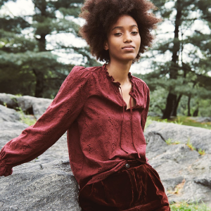 A person in a maroon blouse and dark pants sits on a large rock, with a natural forest background of trees and greenery