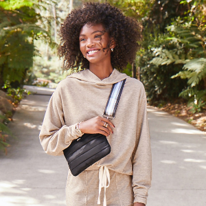 A person wearing a beige hoodie and holding a black bag smiles while walking on a sunlit pathway surrounded by lush, green foliage
