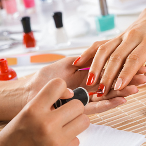 Hands receiving a manicure, nail technician applying red nail polish; various nail polish bottles and tools scattered on the table in a well-lit salon