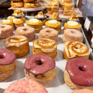 Pastries topped with various icings and garnishes are displayed on a tray in a bakery setting, surrounded by more baked goods on other trays and shelves
