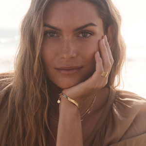 A woman with long hair rests her chin on her hand while wearing gold jewelry, sitting in a sunlit outdoor setting with a blurred background