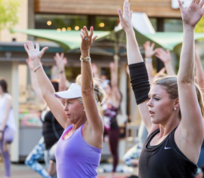 Women exercise, raising arms during a group fitness class, outdoors with others performing the same actions, in front of market stalls and shaded by green umbrellas