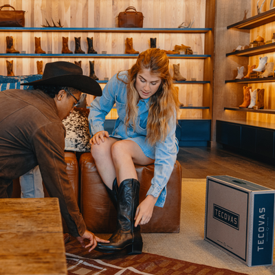 A young woman tries on cowboy boots while seated on a leather bench as a store attendant in a hat assists Rows of boots are displayed on wooden shelves in the background